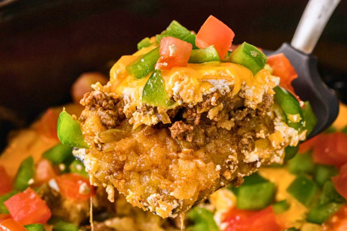 A spoonful of a cheesy beef casserole topped with green peppers and tomatoes, showcasing a hearty mixture of ground beef and rice.