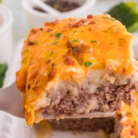 A close-up view of a slice of cheesy meatloaf casserole with a golden top, held by a spatula, showcasing layers of beef, mashed potatoes, and melted cheese.