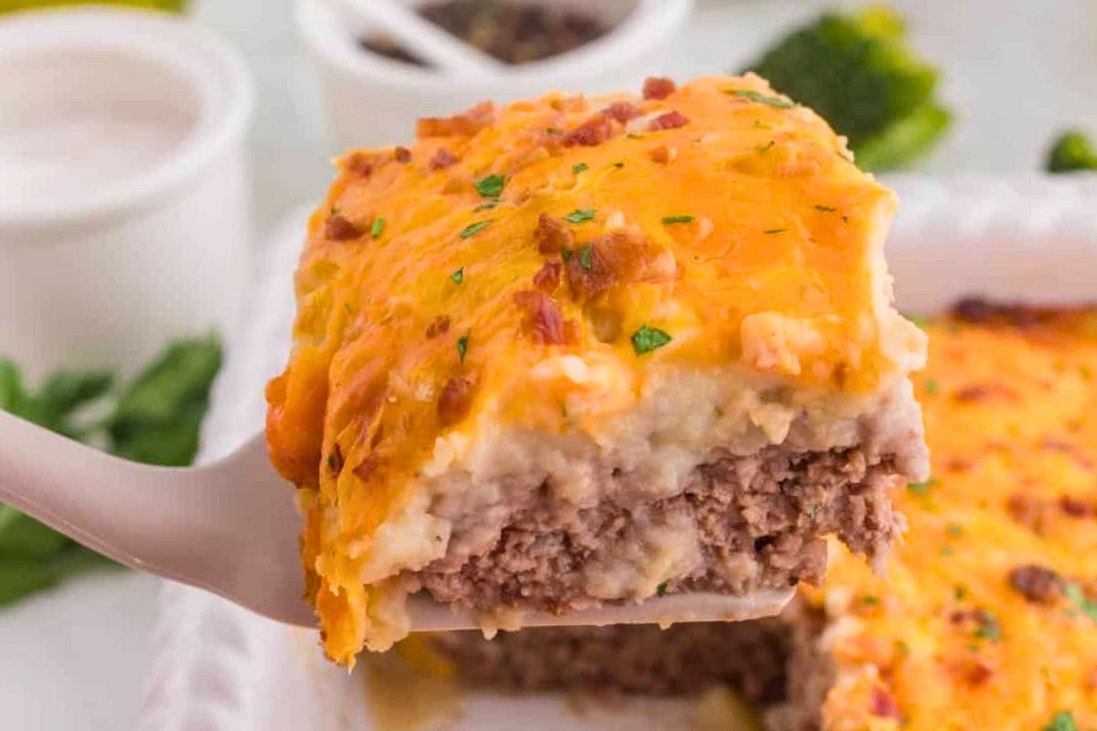 A close-up view of a slice of cheesy meatloaf casserole with a golden top, held by a spatula, showcasing layers of beef, mashed potatoes, and melted cheese.