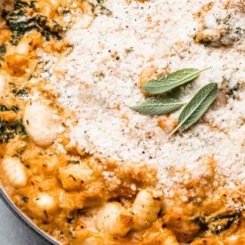 A close-up of a baked dish featuring gnocchi, spinach, and a creamy sauce, topped with grated cheese and fresh sage leaves.