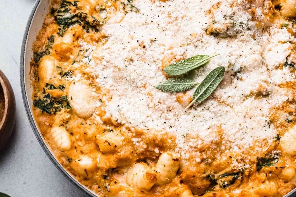 A close-up of a baked dish featuring gnocchi, spinach, and a creamy sauce, topped with grated cheese and fresh sage leaves.