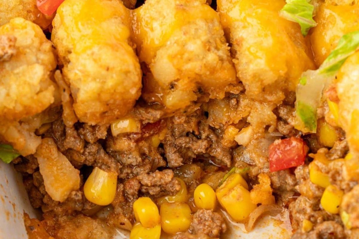 A close-up of a cheesy tater tot casserole with ground beef, corn, and diced tomatoes, showcasing its golden, crispy tater tots on top.
