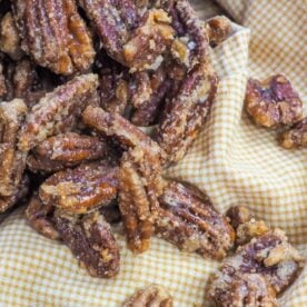 A close-up of sweet, candied pecans spilling from a glass jar onto a yellow checkered cloth.