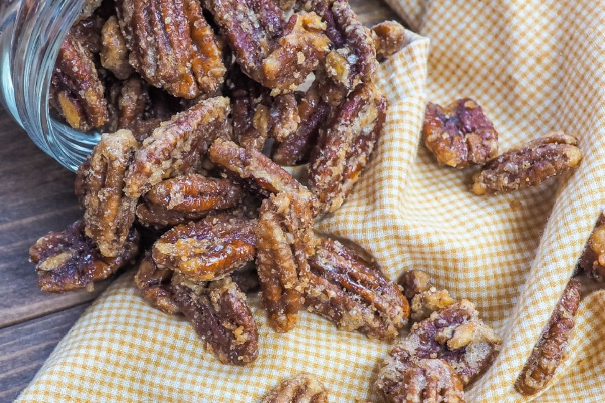A close-up of sweet, candied pecans spilling from a glass jar onto a yellow checkered cloth.