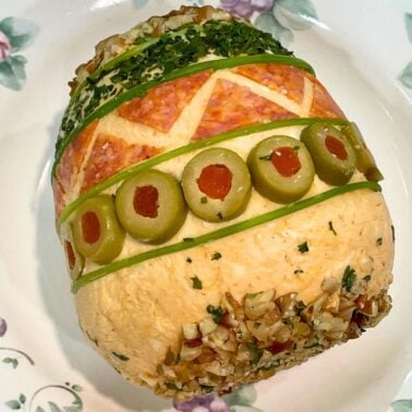 A beautifully arranged cheese ball, adorned with colorful layers of toppings and garnished with olives and herbs, presented on a floral platter.