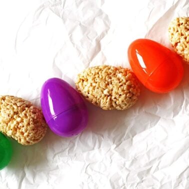 A vibrant display of colorful plastic Easter eggs next to rice crispy treat eggs, set against a crumpled white background.