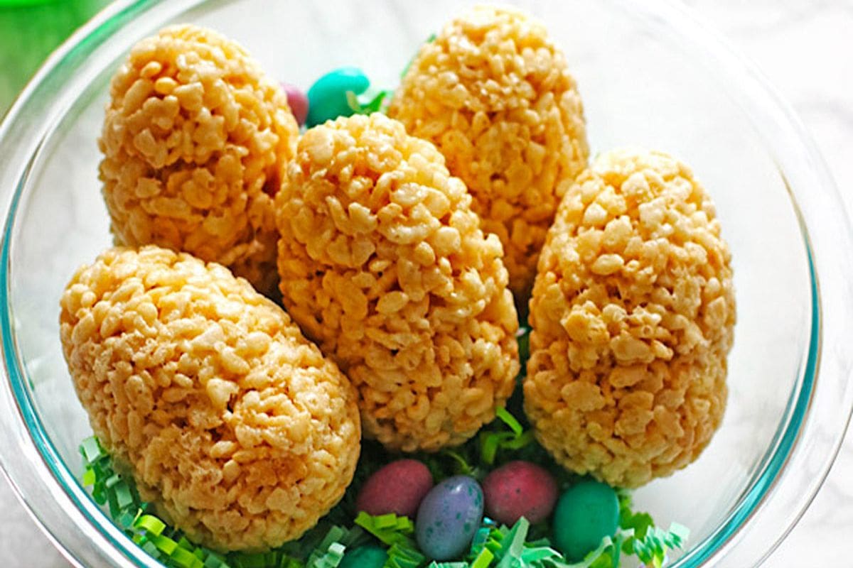 A clear glass bowl filled with rice crispy treats shaped like eggs, surrounded by colorful plastic Easter eggs and green shredded paper.