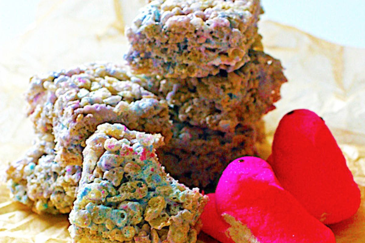 Stack of vibrant rice cereal treats next to pink marshmallow figures on crinkled paper.
