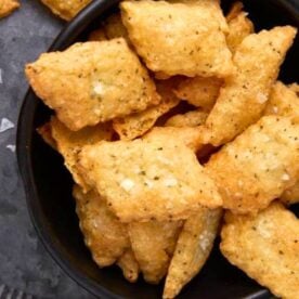 A close-up view of a bowl filled with golden, crispy crackers with herbs and seasoning on a gray surface.
