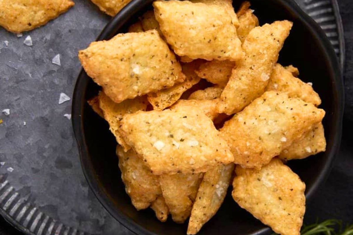 A close-up view of a bowl filled with golden, crispy crackers with herbs and seasoning on a gray surface.