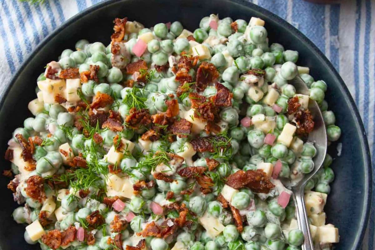 A close-up view of a creamy pea salad featuring green peas, diced cheese, crispy bacon bits, and fresh herbs, served in a black bowl on a striped tablecloth.