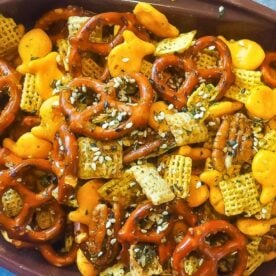 A colorful and crunchy snack mix featuring pretzels, cheese crackers, and cereal, garnished with sesame seeds and seasonings.