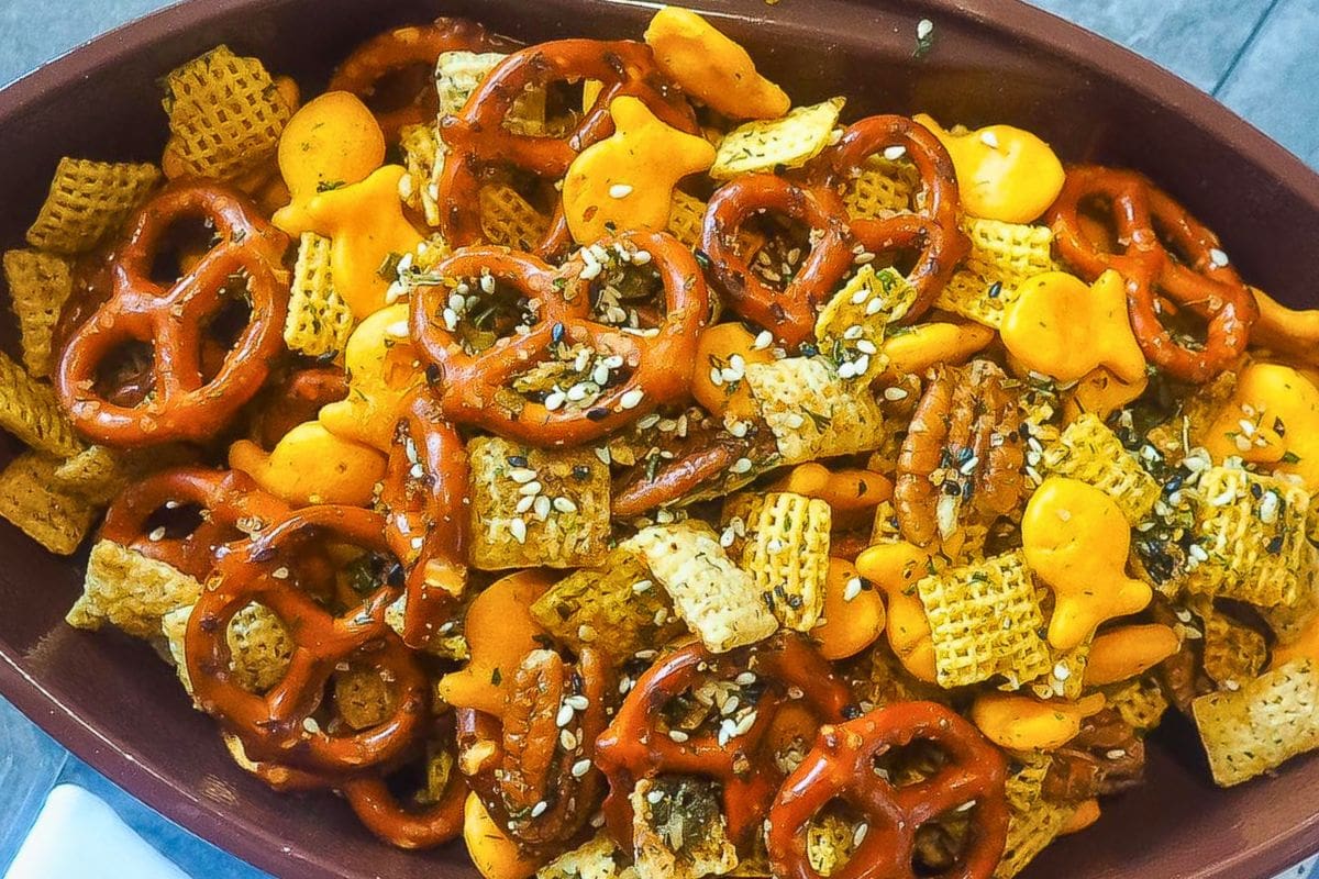 A colorful and crunchy snack mix featuring pretzels, cheese crackers, and cereal, garnished with sesame seeds and seasonings.